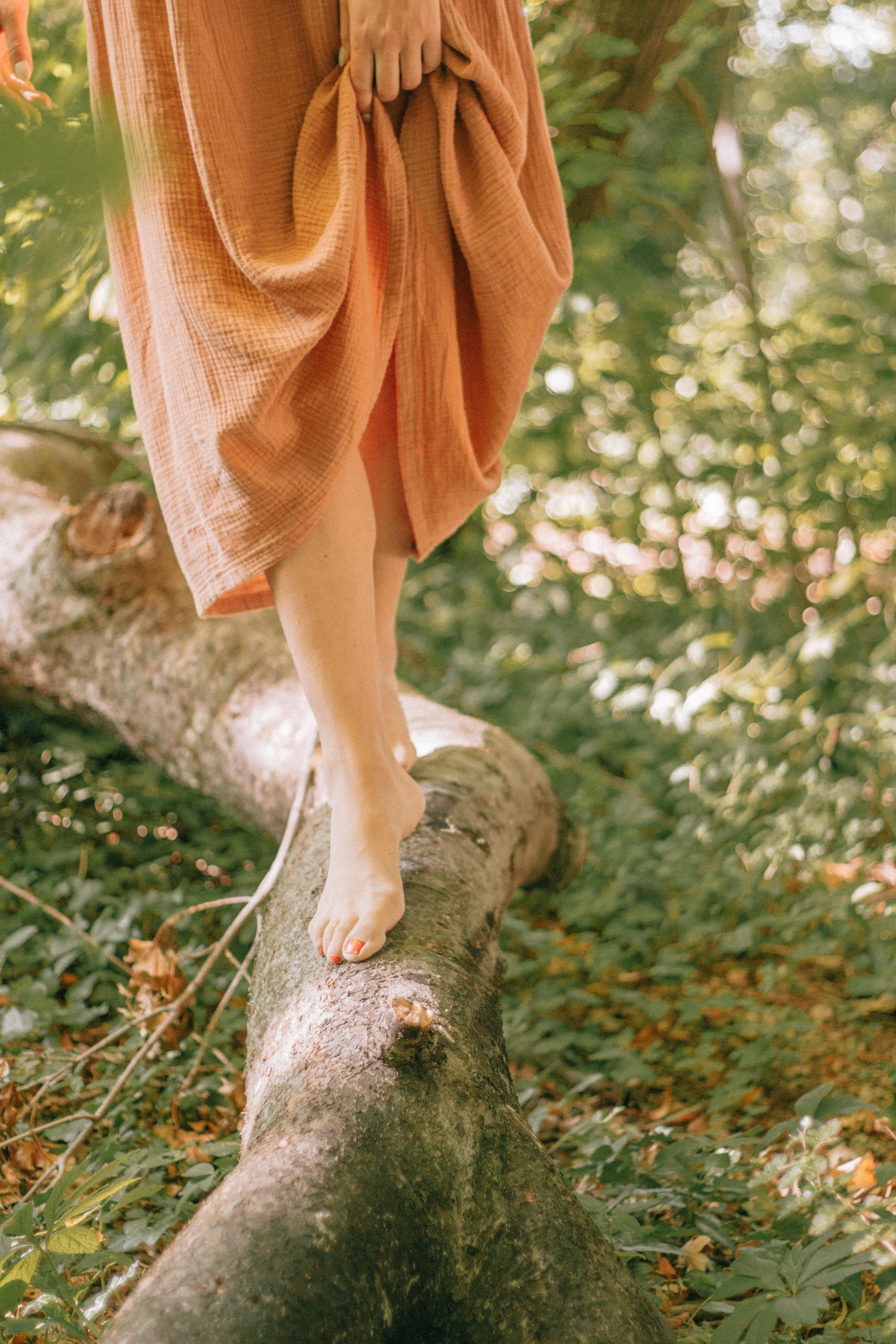 Jambes d'une jeune femme recouvertes d'une jupe couleur orange pale, qui marche en équilibre sur un tronc d'arbre