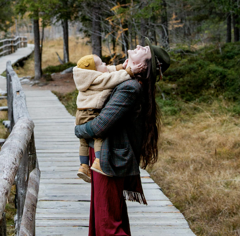 Jeune femme souraint avec un bébé dans les bras dans une ambiance de campagne qui montre la complicité et la joie qui illustre le soin "Bien-être et équilibre postpartum"