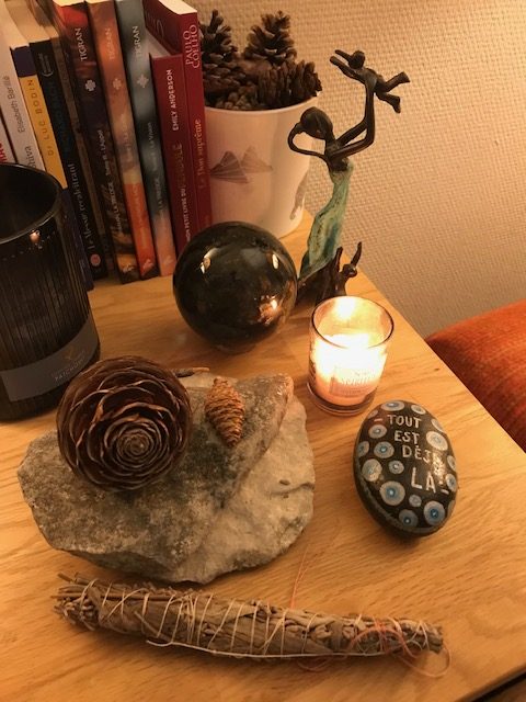 Pierre colorée sur laquelle est écrit: "Tout est déjà là", bougie allumé, sculpture d'une femme qui soulève son enfant. Pierre labradorite ronde. Baton de fumigation de sauge. Le tout posé sur une table en bois. Des livres en arrière plan.