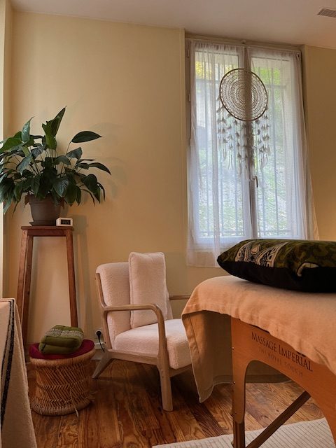 Cabinet de soin avec une table de massage, un fauteuil au coussins clair, un tabouret en osier, une plante verte sur un guéridon haut. Un fenêtre avec des voilages blancs et un grand-attrape re^ve suspendu.