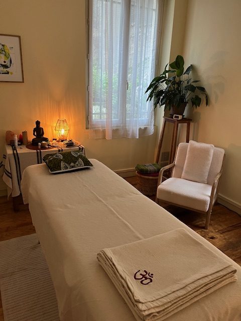 Cabinet de soin avec une table de massage, couverture et fauteuil de couleur claire. Plante verte et petite lampe pour une ambiance chaleureuse.